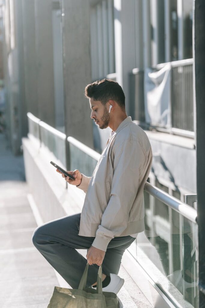 Confident young ethnic guy using smartphone while leaning on fence on street