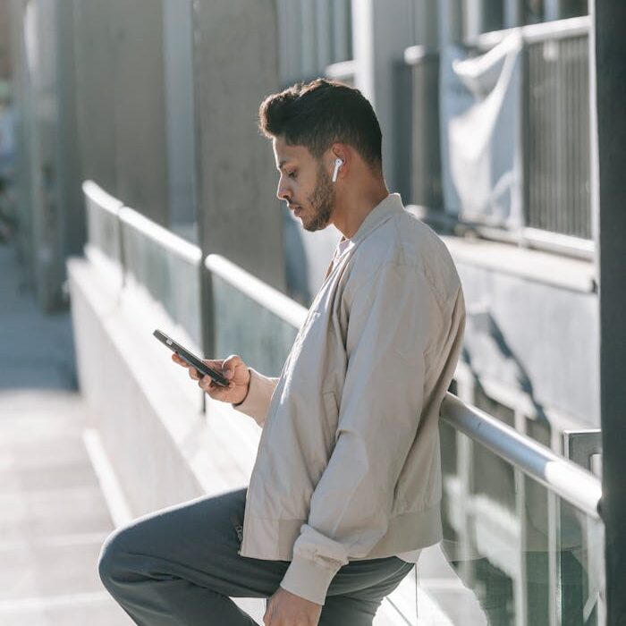 Confident young ethnic guy using smartphone while leaning on fence on street